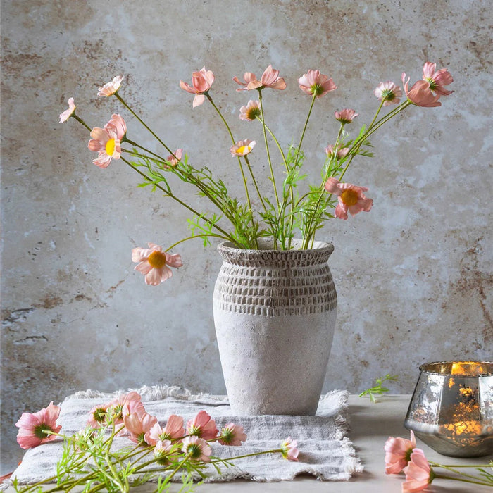 The ABIGAIL AHERN SOFT PINK COSMOS MEADOW arrangement in a textured glass vase sits on a light wood table beside beige and brown tea light holders, creating a soft botanical vibe. A blurred dark chair appears in the background.