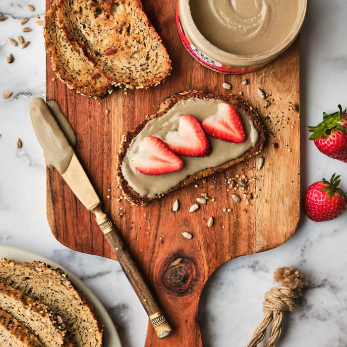 A slice of seeded bread on a wooden board is topped with a creamy vegan spread and three strawberry slices. Nearby are a knife with spread, more bread slices, whole strawberries, and a jar of 88 ACRES Dark Chocolate Sunflower Seed Butter.