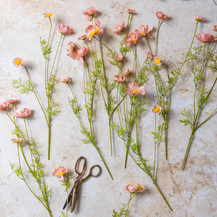 The ABIGAIL AHERN SOFT PINK COSMOS MEADOW arrangement in a textured glass vase sits on a light wood table beside beige and brown tea light holders, creating a soft botanical vibe. A blurred dark chair appears in the background.