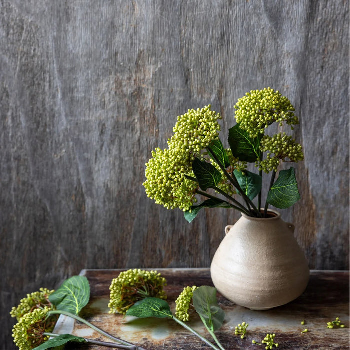 The ABIGAIL AHERN - VIBURNUM OLIVE by ABIGAIL AHERN features a single long artificial stem with two clusters of small yellow-green buds and a large, textured green leaf set against a plain white background.