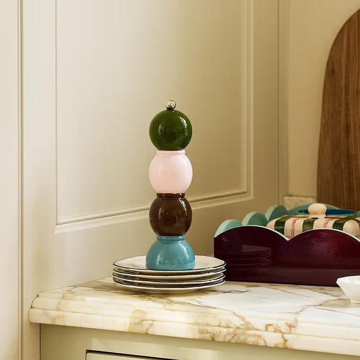 Three vibrant ADDISON ROSS LONDON Bobbin Salt or Pepper Mills (24cm) from ADDISON ROSS are stacked on a kitchen counter near wooden cabinets, with jars of dry food visible in the background.