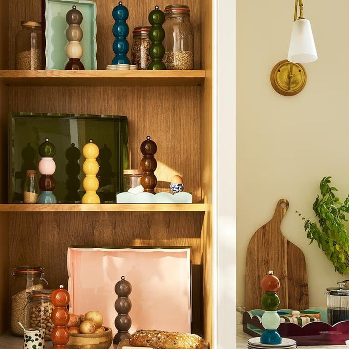 Three vibrant ADDISON ROSS LONDON Bobbin Salt or Pepper Mills (24cm) from ADDISON ROSS are stacked on a kitchen counter near wooden cabinets, with jars of dry food visible in the background.