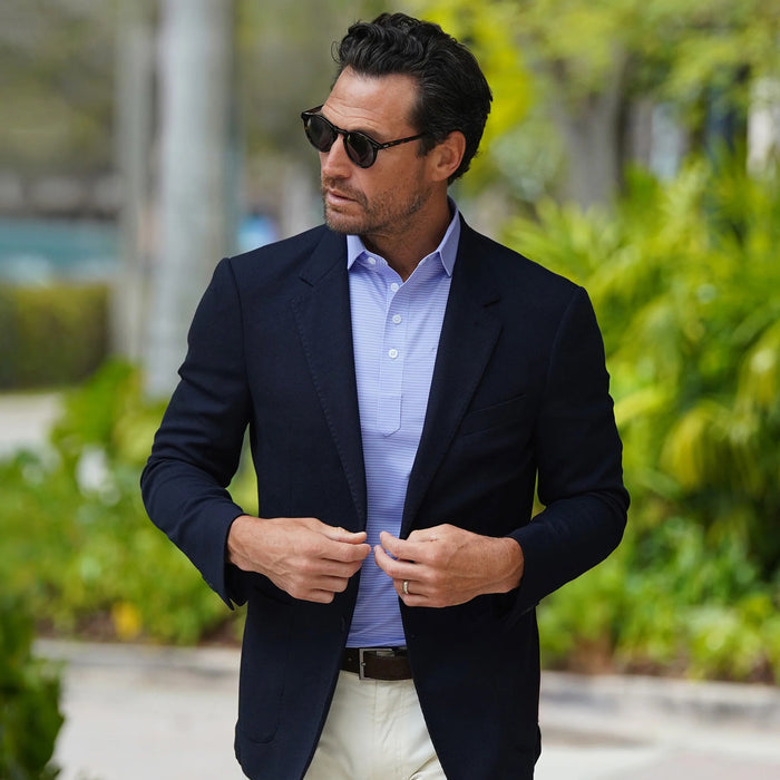 A man wearing sunglasses, a navy blazer, beige pants, and the COLLARS AND CO - Featherweight Flex Polo in Brookline Lavender with Turquoise Stripe stands outdoors in a leafy area, looking to the side and buttoning his jacket.