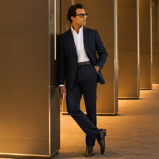Close-up of a person wearing COLLARS AND CO’s Maverick Performance Pants in Black, paired with a dark suit jacket, white shirt, and brown belt. The open jacket reveals the inner lining and label. Light brick background.
