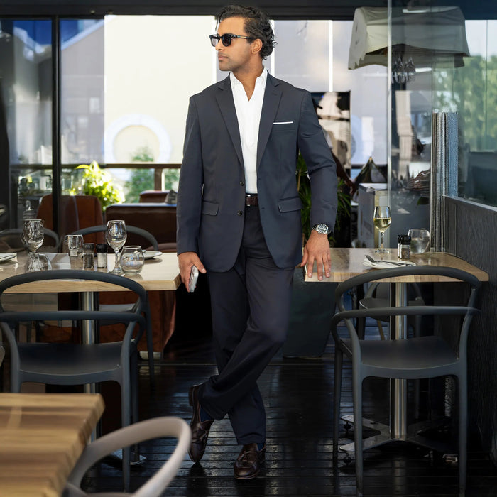 Close-up of a person wearing COLLARS AND CO’s Maverick Performance Pants in Black, paired with a dark suit jacket, white shirt, and brown belt. The open jacket reveals the inner lining and label. Light brick background.