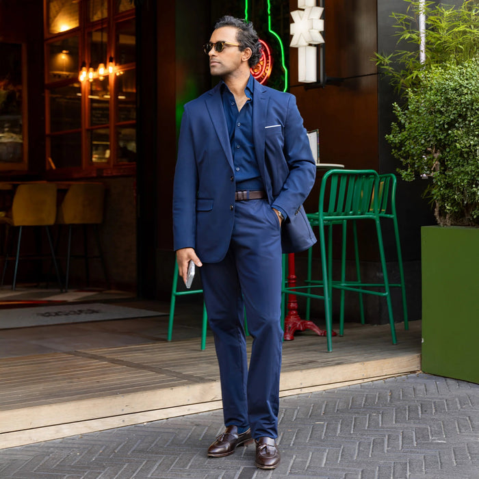 A man in COLLARS AND CO Maverick Performance Pants in Navy, a dark shirt, and sunglasses stands confidently outside a restaurant with green chairs and lush plants nearby, holding his phone.
