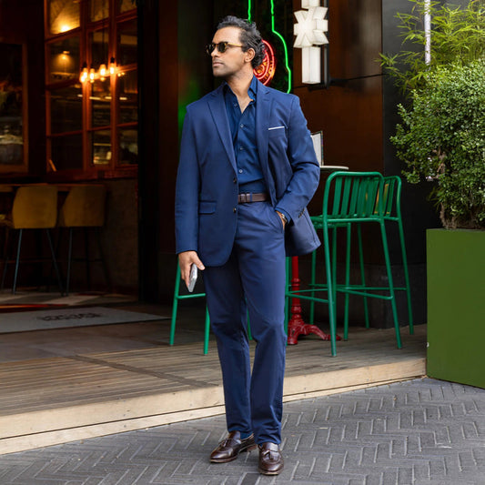 A man in COLLARS AND CO Maverick Performance Pants in Navy, a dark shirt, and sunglasses stands confidently outside a restaurant with green chairs and lush plants nearby, holding his phone.