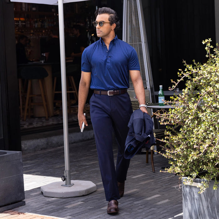 A man in COLLARS AND CO Maverick Performance Pants in Navy, a dark shirt, and sunglasses stands confidently outside a restaurant with green chairs and lush plants nearby, holding his phone.