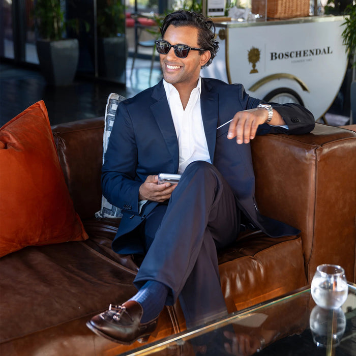A man in COLLARS AND CO Maverick Performance Pants in Navy, a dark shirt, and sunglasses stands confidently outside a restaurant with green chairs and lush plants nearby, holding his phone.