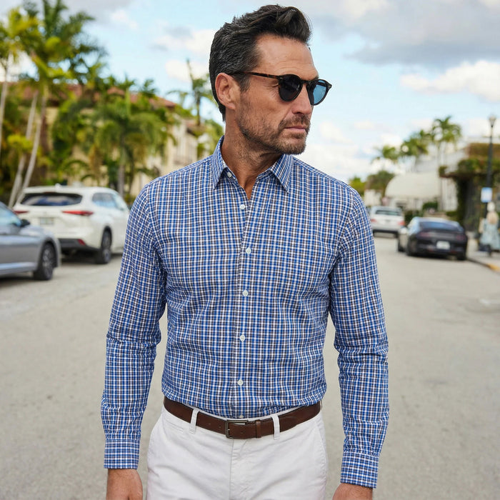 A man wearing sunglasses and the COLLARS AND CO - Semi Spread Collar Apex Performance Dress Shirt in Royal and Brown Tattersall stands confidently on a sunny, palm-lined street.