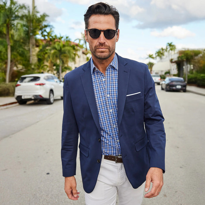 A man wearing sunglasses and the COLLARS AND CO - Semi Spread Collar Apex Performance Dress Shirt in Royal and Brown Tattersall stands confidently on a sunny, palm-lined street.