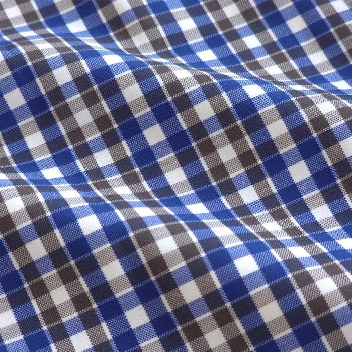 A man wearing sunglasses and the COLLARS AND CO - Semi Spread Collar Apex Performance Dress Shirt in Royal and Brown Tattersall stands confidently on a sunny, palm-lined street.