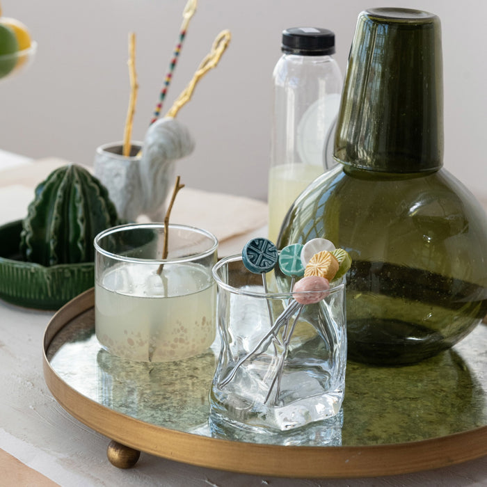 A round tray displays two CREATIVE COOP HAND-BLOWN ROUND CONFETTI DRINKING GLASSES with beverages, decorative stirrers, a green bottle, lemonade bottle, and a small cactus ornament on a tabletop with light linens.