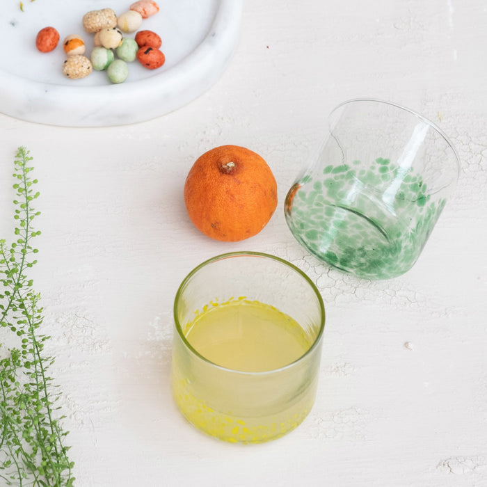A round tray displays two CREATIVE COOP HAND-BLOWN ROUND CONFETTI DRINKING GLASSES with beverages, decorative stirrers, a green bottle, lemonade bottle, and a small cactus ornament on a tabletop with light linens.