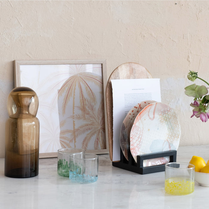 A round tray displays two CREATIVE COOP HAND-BLOWN ROUND CONFETTI DRINKING GLASSES with beverages, decorative stirrers, a green bottle, lemonade bottle, and a small cactus ornament on a tabletop with light linens.