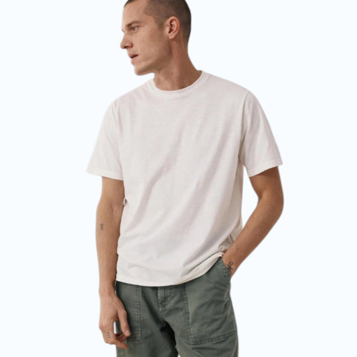 Wearing the HARTFORD - SLUB COTTON CREWNECK T-SHIRT IN OFF WHITE, a short-haired person in green pants stands against a light background with hands loosely clasped, small wrist tattoos visible.