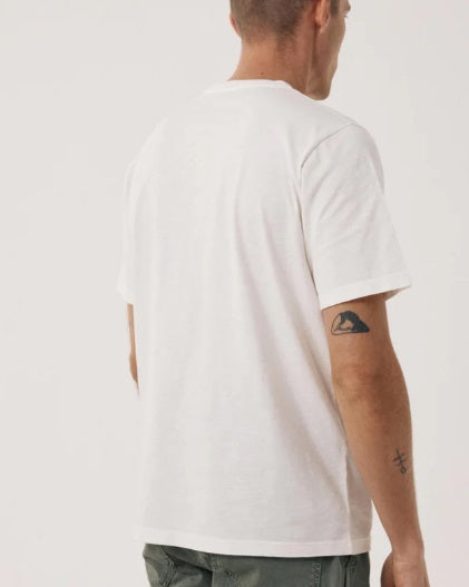 Wearing the HARTFORD - SLUB COTTON CREWNECK T-SHIRT IN OFF WHITE, a short-haired person in green pants stands against a light background with hands loosely clasped, small wrist tattoos visible.