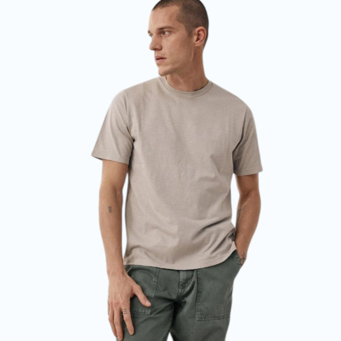 A man with short hair wears the HARTFORD Slub Cotton Crewneck T-shirt in Sand and green pants, standing with one hand in his pocket and looking to the side against a light background.