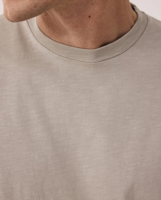 A man with short hair wears the HARTFORD Slub Cotton Crewneck T-shirt in Sand and green pants, standing with one hand in his pocket and looking to the side against a light background.
