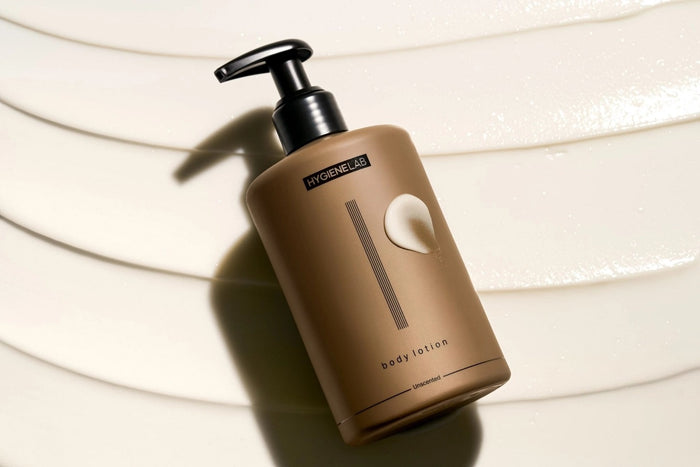 A four-part image showcases the HYGIENELAB - BODY LOTION, UNSCENTED, spotlighting key ingredients Tocopherol, Squalane, and Aloe Barbadensis along with their skin benefits. The composition features a “Clean Non-Toxic Formula” bottle from HYGIENELAB bearing a health certification badge. This dermatologist-approved body lotion is also EWG Verified, ensuring your peace of mind.