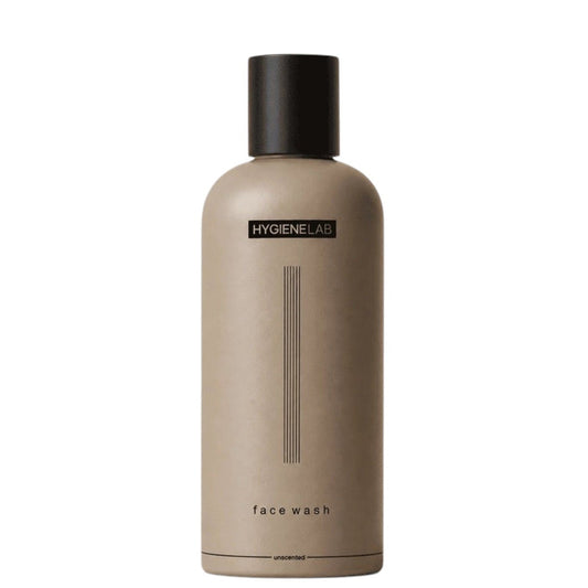 A person dispenses HYGIENELAB - FACE WASH, UNSCENTED from a brown bottle onto their open palm against a neutral background.