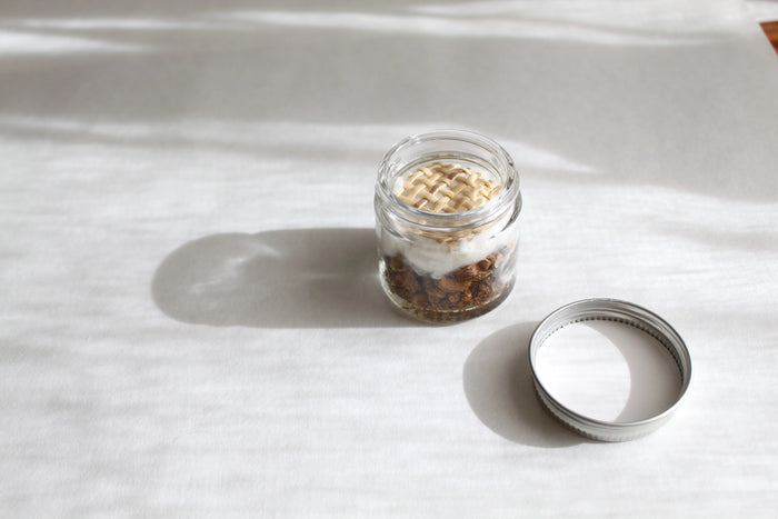 A compact glass jar with a white lid, labeled “MADISON AND GREEN - WHILE DRIVING DISTRESS RELIEF AROMATHERAPY INHALER” by MADISON GREEN, filled with a blend of dried botanicals and essential oils designed for on-the-go stress relief.