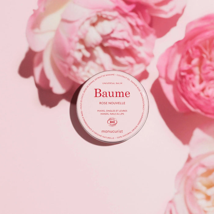 A round, pink tin of MANUCURIST - ROSE BALM COSMOS ORGANIC NATURAL NOURISHING BALM from MANUCURIST INC with the lid slightly ajar to reveal the balm inside. The label notes its versatility for hands, nails, and lips—making it an ideal nourishing cuticle balm and lip repair treatment that easily integrates into your natural skincare routine.