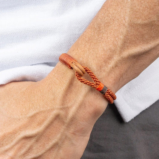 A close-up of a person's wrist wearing the MAKARLA MEN'S NAUTICAL ROPE BRACELET IN CHOCOLATE, featuring braided rope and a wooden clasp, set against a white fabric background.