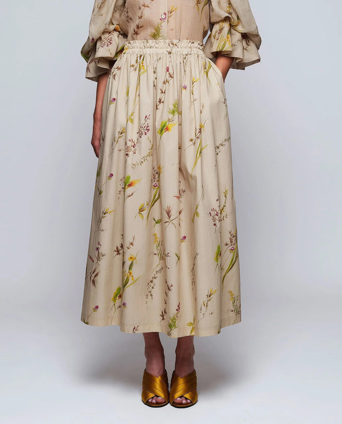 A woman stands against a plain background wearing the MIRTO 1956 Beige Floral Print Midi Skirt, paired with puffed three-quarter sleeves, a wide tan belt, and brown open-toe heels. Her hair is styled in a high bun with a decorative accessory.