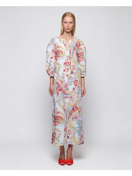 A woman with long blonde hair is seen from the back, wearing the MIRTO 1956 Paisley Print Long Sleeve Dress; its vibrant pattern and puffy sleeves flow to ankle length, revealing bright red heeled shoes against a white background.