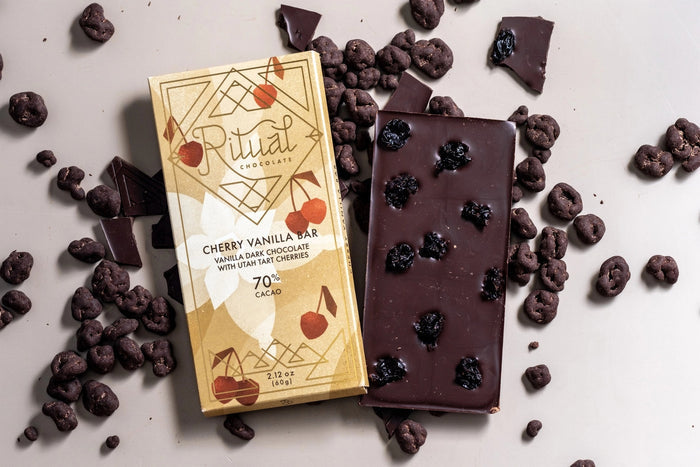 A beige RITUAL CHOCOLATE - CHERRY VANILLA DARK CHOCOLATE BAR 70% wrapper features geometric patterns, cherry art, and the text: "Vanilla Dark Chocolate with tart cherries, 70% Cacao, 2.12 oz (60g).