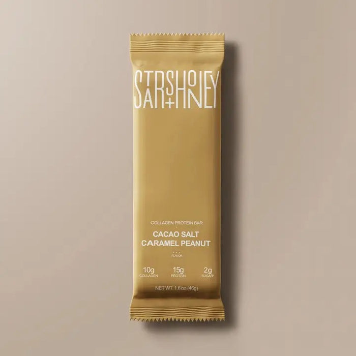 A gold protein bar wrapper labeled “STARS+HONEY” featuring the flavor “Cacao Salt Caramel Peanut” is placed on a beige background, highlighting 10g collagen, 15g protein, and just 2g sugar as a low sugar protein bar option.