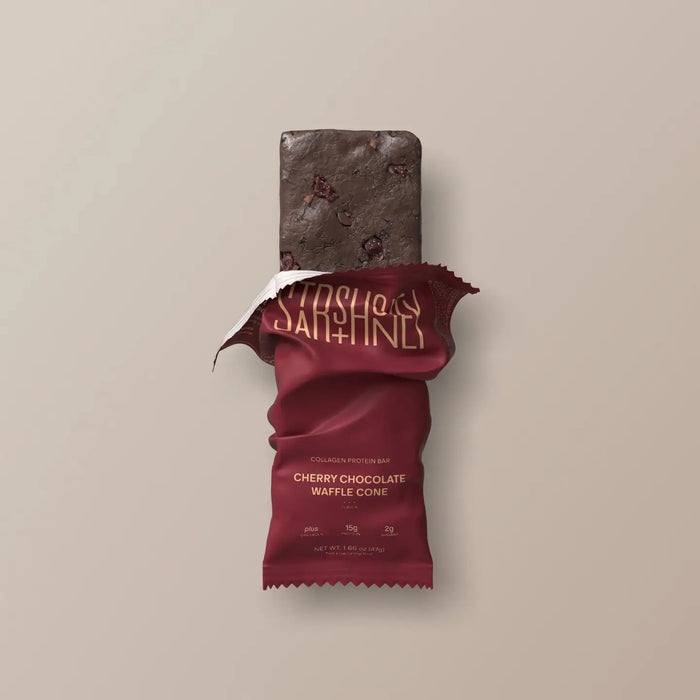 A maroon protein bar wrapper labeled “STARS+HONEY” displays the flavor “CHERRY CHOCOLATE WAFFLE CONE” with nutrition highlights: 8g collagen, 15g protein, and 2g sugar, all set against a beige background. This indulgent treat puts the rich chocolate and tart cherry combination front and center.